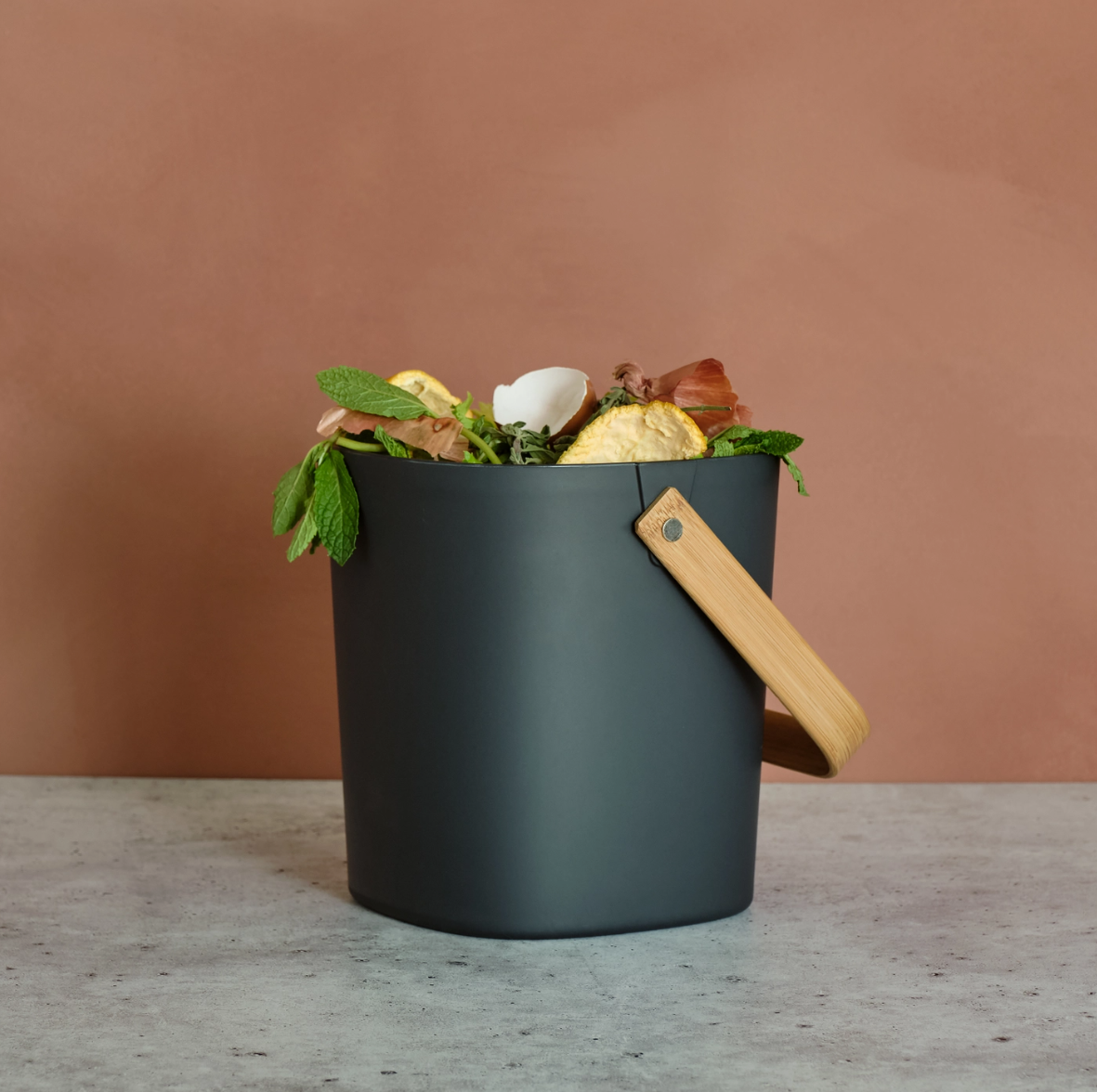 Black compost bin with a wooden handle on a light wood surface against a brown background