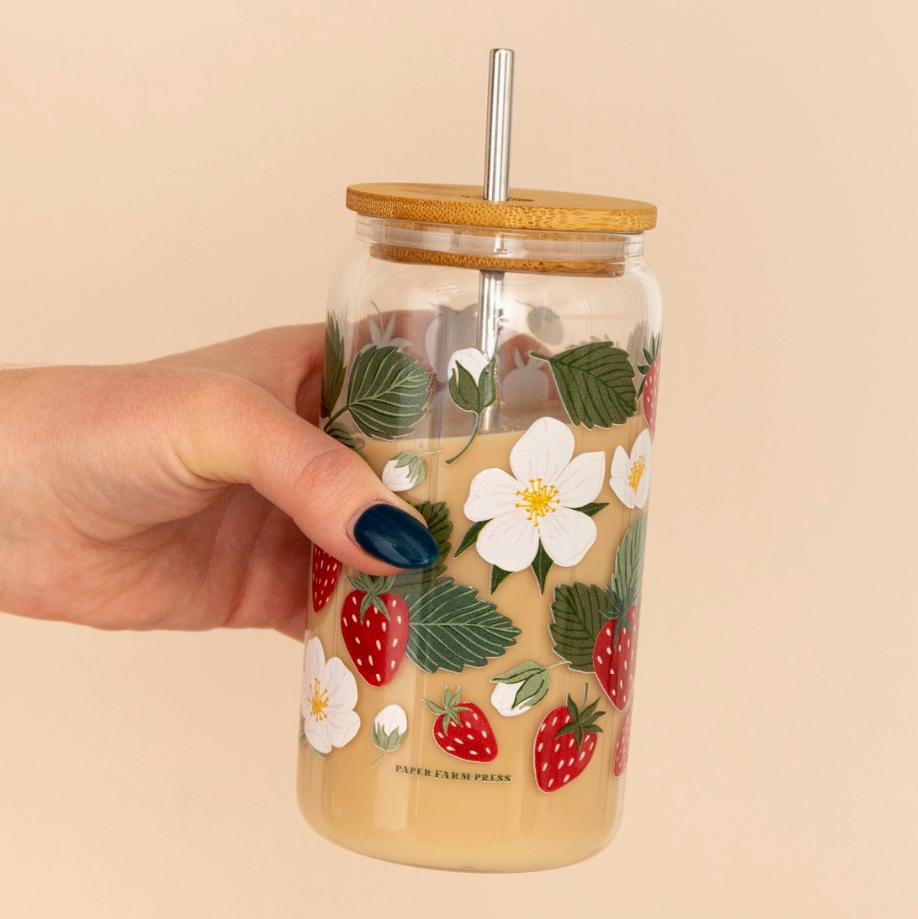Hand holding a glass tumbler with floral and strawberry design on a beige background
