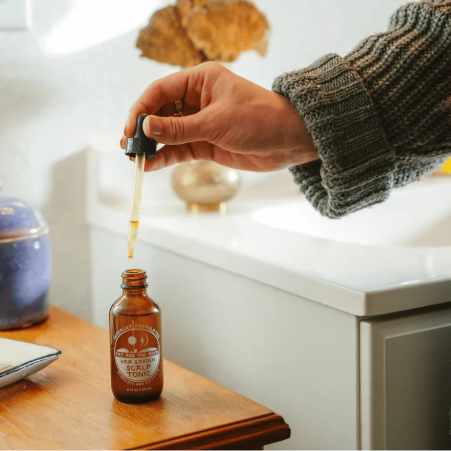 Person using a dropper to add liquid from a bottle labeled 'Scalp Tonic' on a wooden surface.