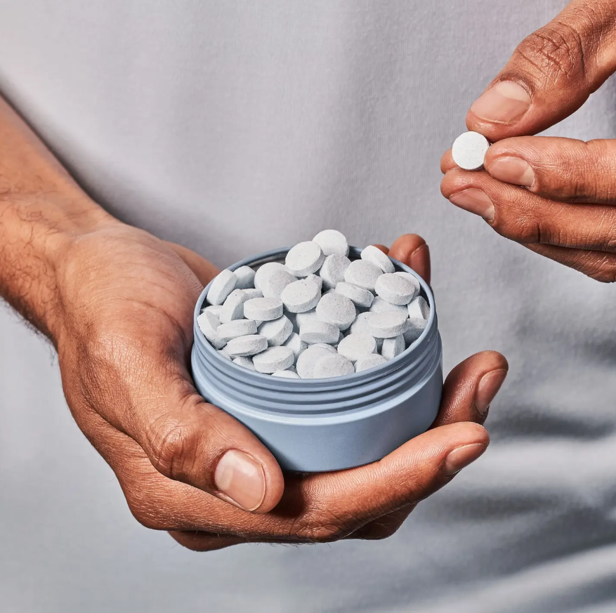 A close-up of a person holding a small round container filled with white round tablets, while another hand holds a single tablet above it against a plain light gray background.