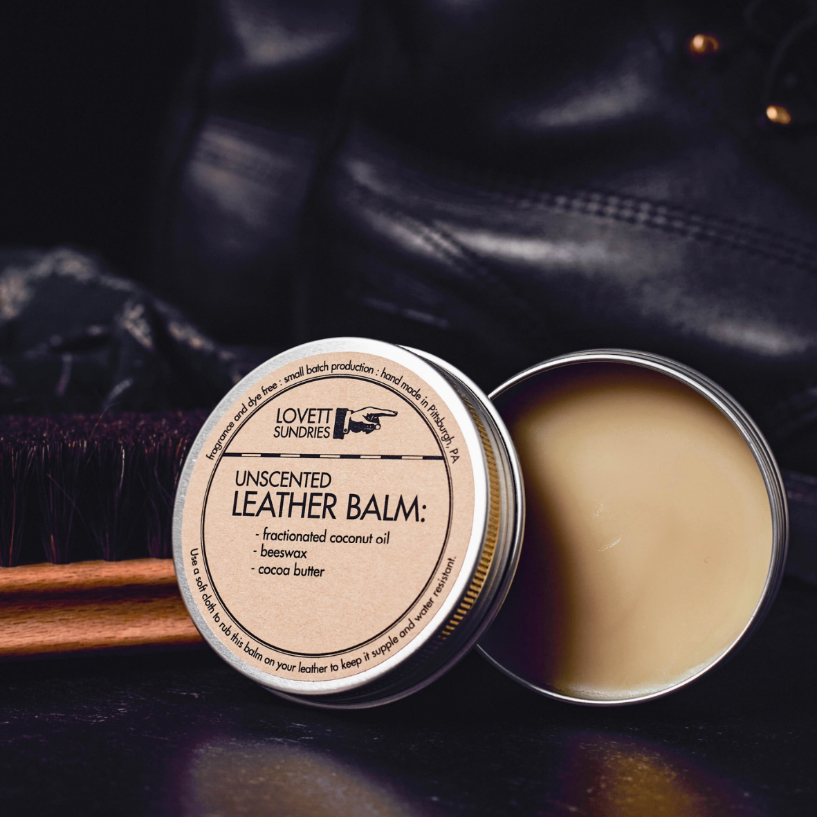 Unscented leather balm in a metal container with a wooden applicator on a dark background.