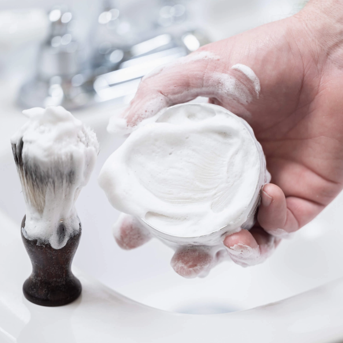 Hand holding a glass filled with white shaving cream next to a black-handled shaving brush.