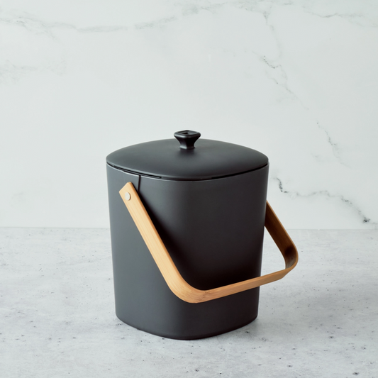 Black compost bin with a wooden handle on a light wood surface against a brown background