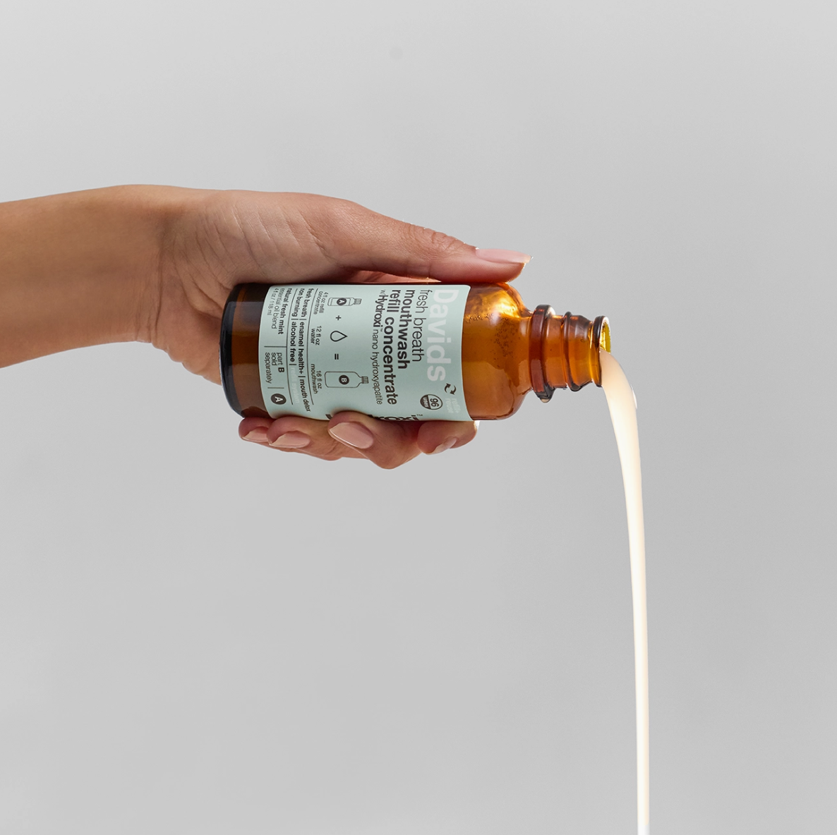 Hand holding a brown bottle with a white label, pouring a liquid onto a gray background