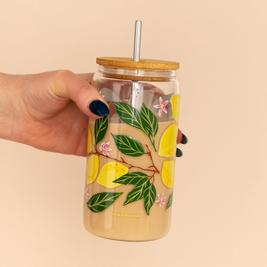 Hand holding a glass tumbler with lemon and leaf design on a beige background