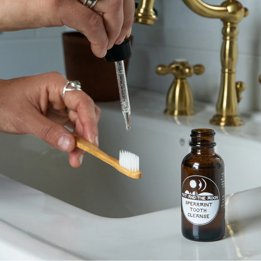 Person using a dropper to apply liquid to a wooden toothbrush with a bottle of Spearmint Tooth Cleanse in the foreground.