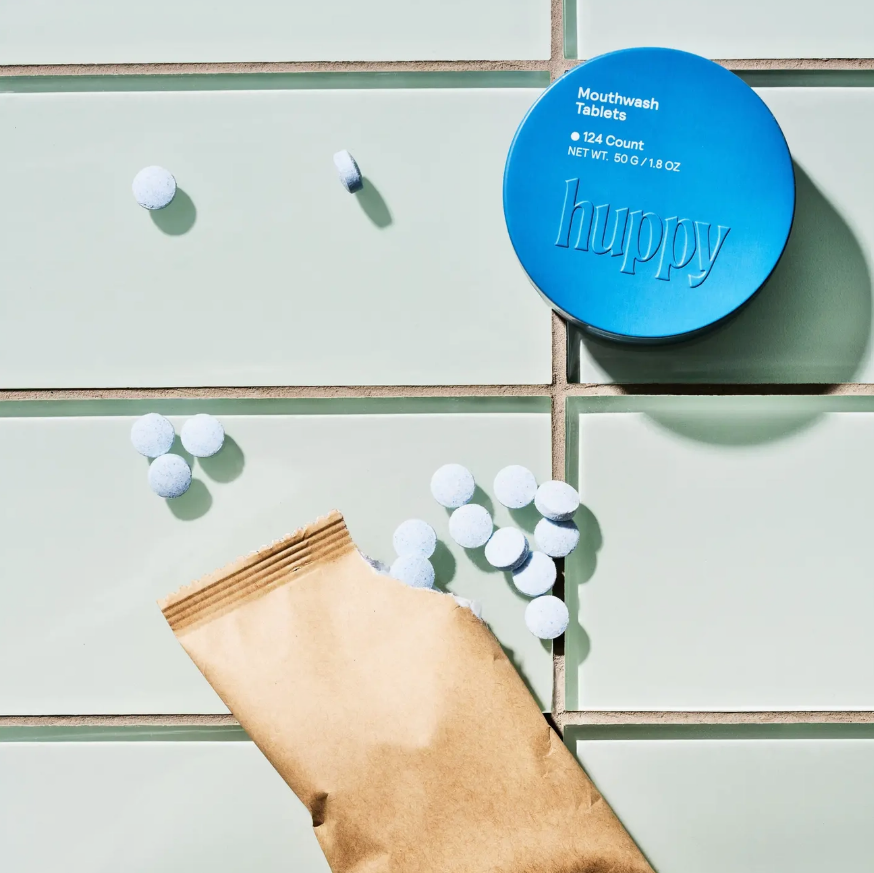 A top-down styled product shot showing a round blue container labeled 'happy' with mouthwash tablets, scattered white tablets, a brown paper pouch, and pale mint-green tiled background.