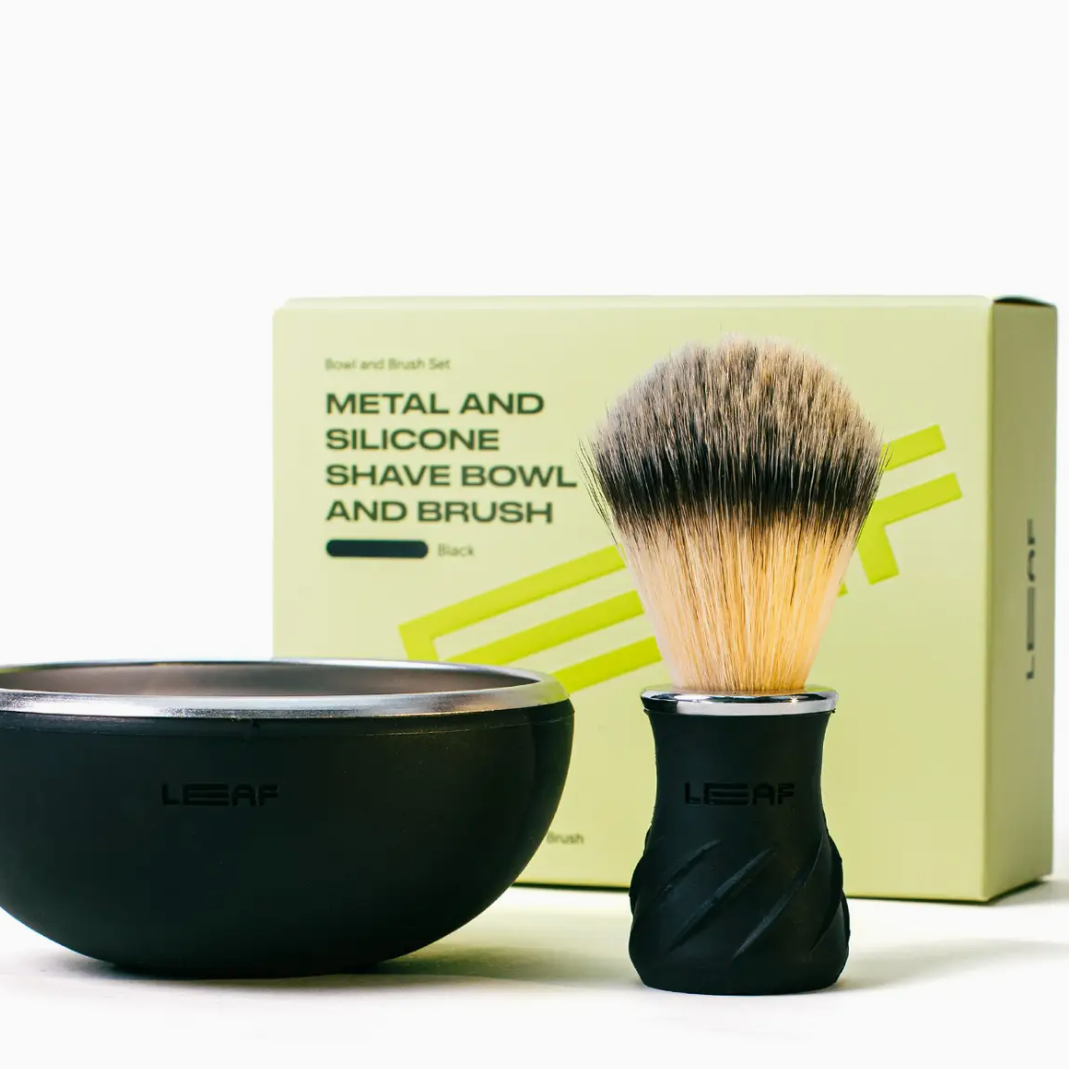 A LEAF shaving set featuring a matte black bowl with a brushed silver interior, a black shaving brush with a dual-toned bristle head, and a light yellow-green product box in the background.