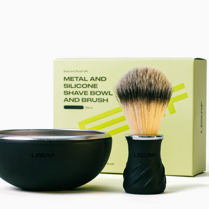 A LEAF shaving set featuring a matte black bowl with a brushed silver interior, a black shaving brush with a dual-toned bristle head, and a light yellow-green product box in the background.