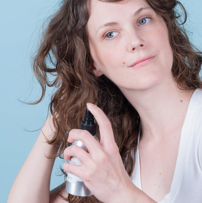 A woman with wavy brown hair holds a silver and black spray bottle against her hair, set against a solid light blue background.